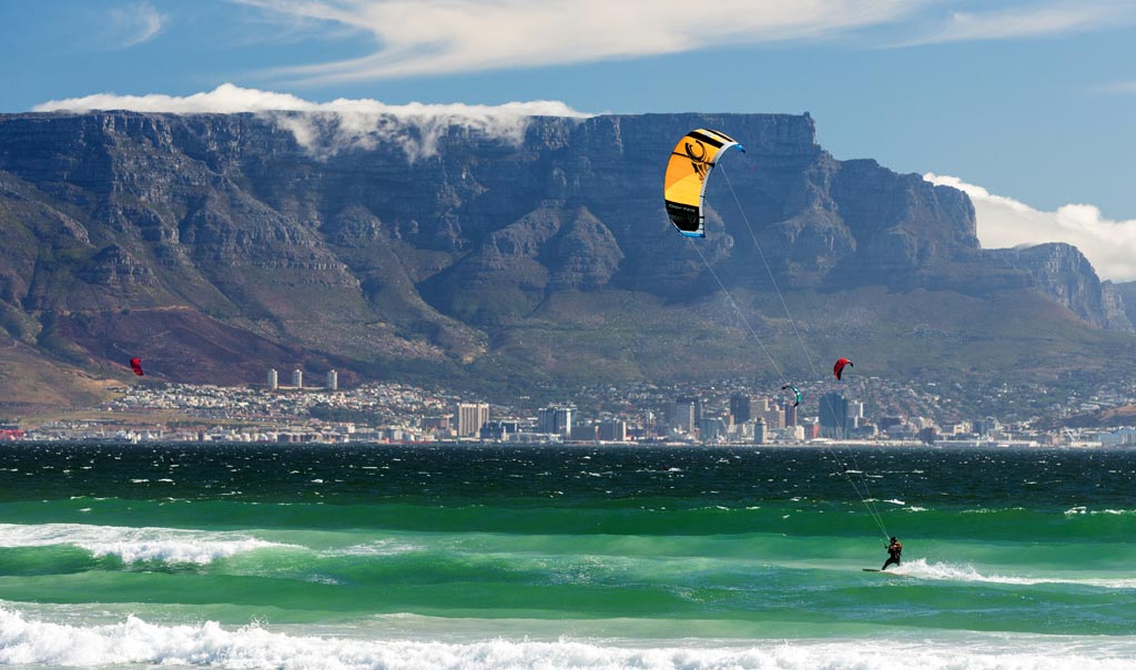 Wide sandy beach in Cape Town, South Africa, with multiple colorful kitesurfers in action and Table Mountain in the background.