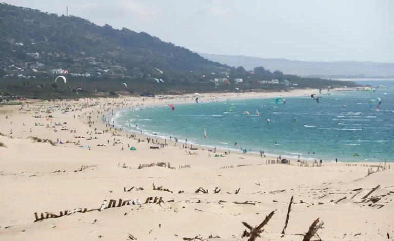 Wide sandy beach in Tarifa Valdevaqueros, Spain, with multiple colorful kitesurfers in action.